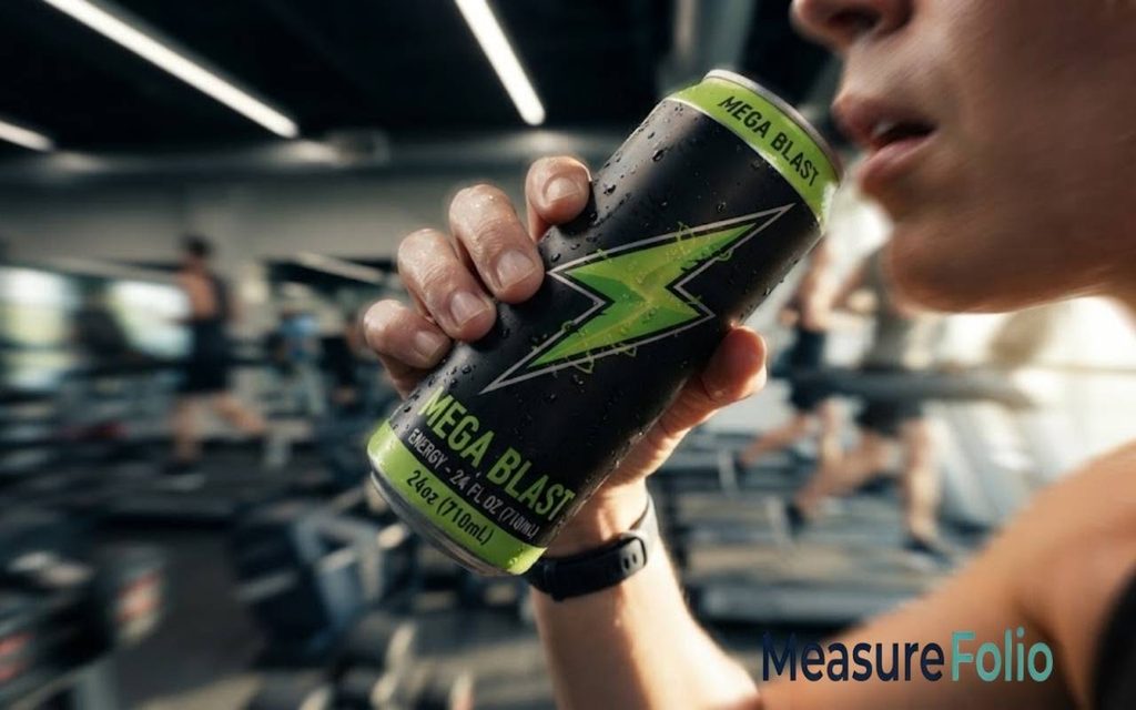 A person holds a black and green "Mega Blast" energy drink can to take a sip.
The background shows a motion-blurred gym with people working out on treadmills.
