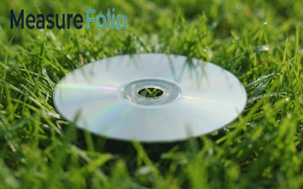 A single silver compact disc rests flat on a bed of lush, green grass.
The reflective data surface catches the sunlight, and morning dew is visible on nearby blades.